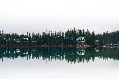 Still lake reflection of cabins surrounded by dense forest, on an overcast da Stock Photos