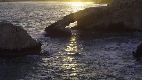 Stone arch in a waving sea. Setting sun shine through the hole in the rock Видео 76988882