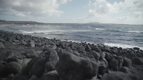 Stone beach with sea waves, cloudy and windy day, volcano in the distance Stock Footage 235677241