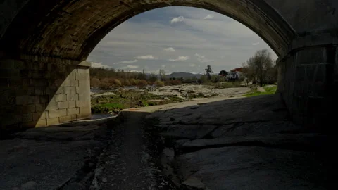 Stone bridge arch framing sunlit river with rocky banks Stock Footage 335461806