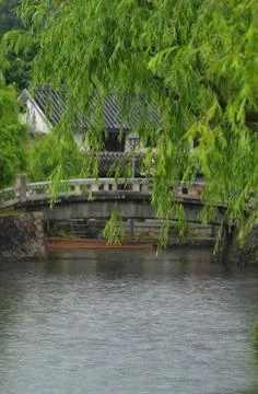 A stone bridge, framed by a willow tree, arches over a river. Stock Photos