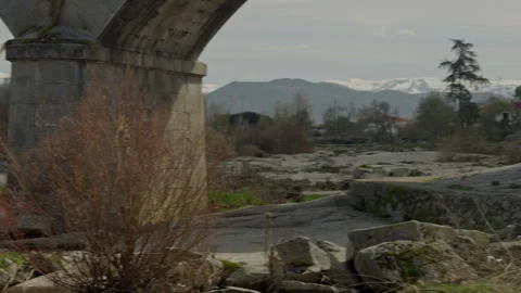 Stone bridge standing over a dry riverbed with green bushes and snow capped.. Stock Footage 335461532