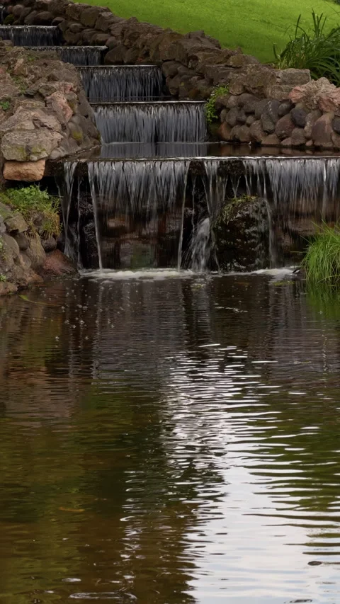 Stone garden waterfall flowing into calm pond with lush green surroundings and Stock Footage 345456430