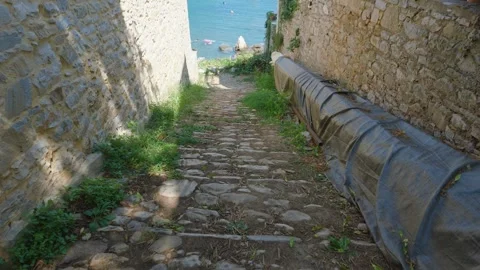 A stone-paved pathway descends between old stone walls toward the turquoise sea Stock Footage 346667043
