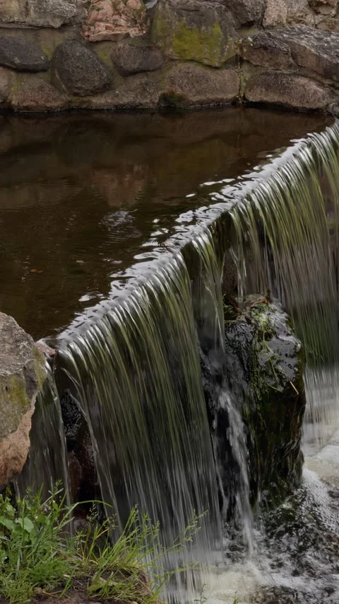 Stone weir waterfall with flowing water into calm pond beside mossy rocks and Stock Footage 345455458
