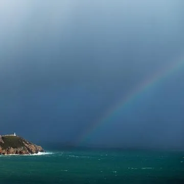 Storm clouds with rainbows approach a lighthouse on the rocky coast Стоковые фото