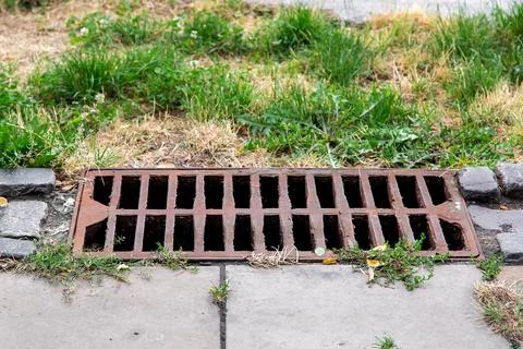 Storm hatch sewage with a rusty grill on the edge of pedestrian stone walkw. Stock Photos