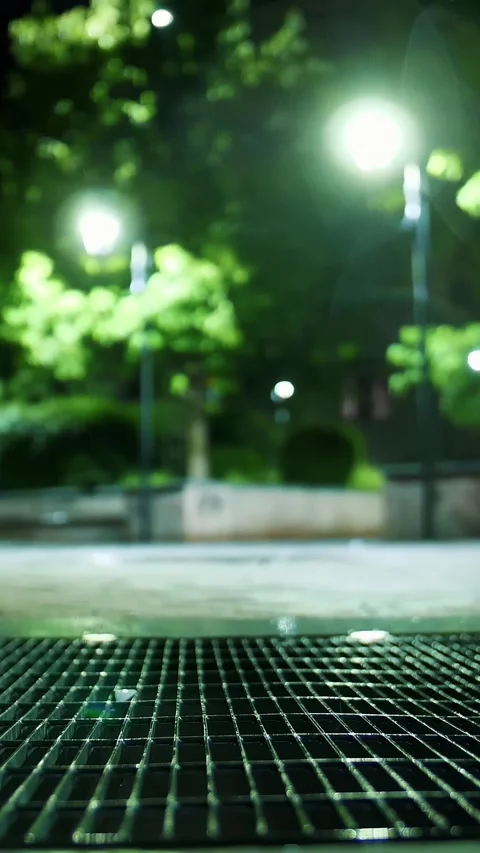 Storm sewer. Light rainfall. Puddle with lights reflection on city sidewalk. Stock Footage 311645109