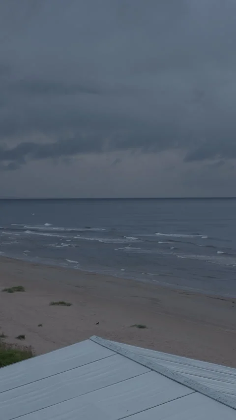 Stormy seaside landscape with empty beach, calm waves, dark clouds, and rooftop Stock Footage 345530498