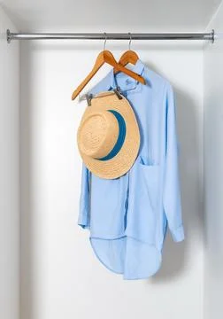 Straw hat and blue dress-shirt hanging in white wardrobe in white closet. Stock Photos