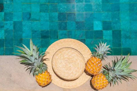 Straw hat and three pineapples by the refreshing pool, viewed from above. Stock Photos