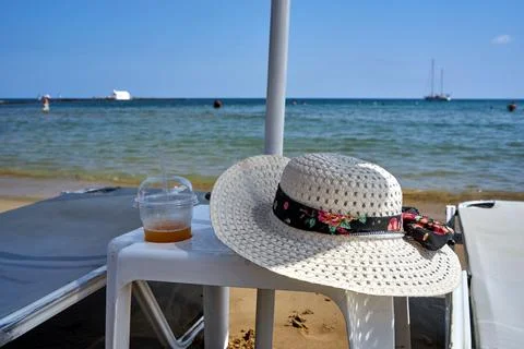 Straw hat, mug with cold drink and sunbathing bed on the sandy beach Stock Photos