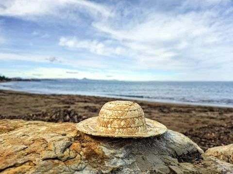Straw hat rests on a rock by the beach during bright daylight with clouds i.. Stock Photos