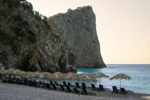 Straw sun umbrellas and empty beach chairs on a pebble shore under towering m Stock Photos