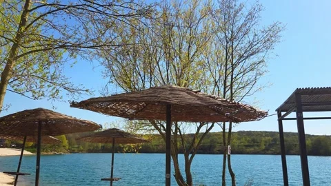 Straw umbrellas on an empty beach near a calm blue lake under a clear sky Stock Footage 337654164