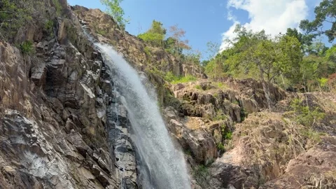 A stream of water falls down a rocky waterfall slope surrounded by vegetation 스톡 동영상 335759893