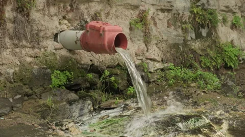 Stream of water flows through pipe in stone wall to street Stock Footage 292311772