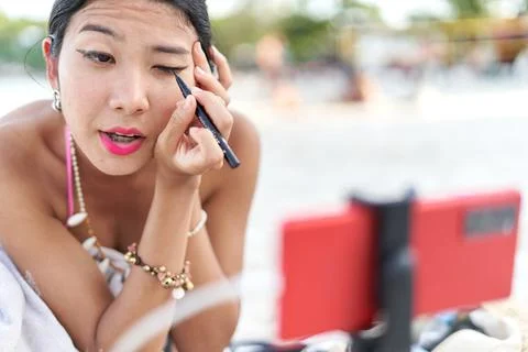 Streamer having her eyes lined using the mobile as mirror on a beach Stock Photos