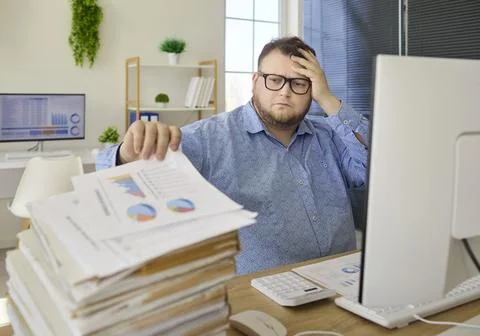 Stressed accountant at desk with paperwork stack and charts on computer screen Stock Photos