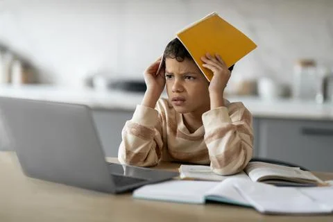 Stressed Black Boy Using Laptop And Covering Head With Workbook At Home Foto stock