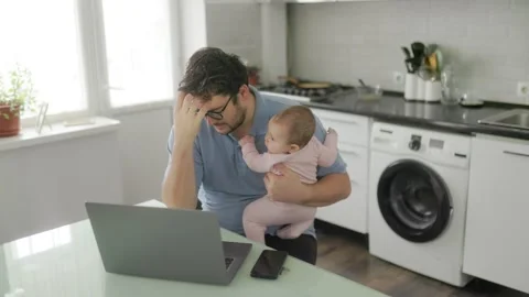 Stressed dad multitasking with infant and laptop in modern kitchen Stock Footage 325323124