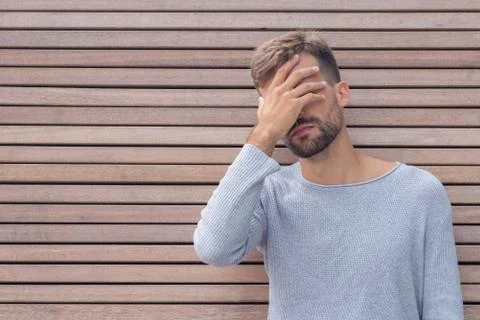 Stressed disappointed young guy making facepalm gesture with hand. Wooden wal Stock Photos