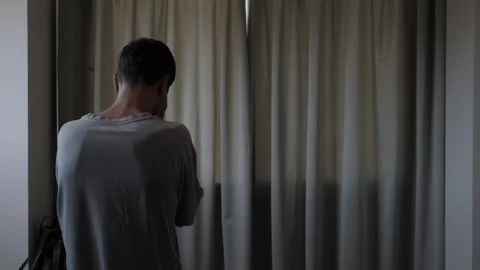 Stressed man standing alone by window during difficult emotional moment Stockbeeldmateriaal 347303175