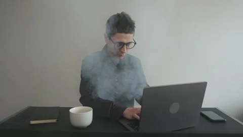 Stressed man working at laptop surrounded by smoke symbolizing burnout and 스톡 동영상 309204979