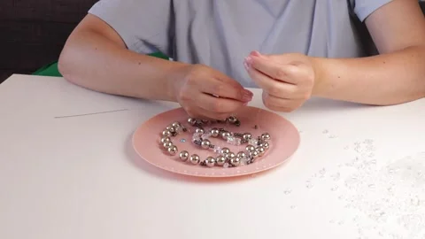 Stringing shiny beads onto a clear fishing line on a pink plate under soft .. Video stock 318671745