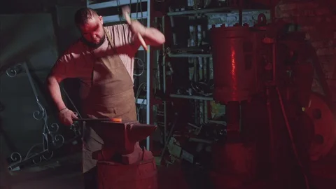 Strong bearded blacksmith is hitting the hammer on a red hot rod at the anvil Video stock 108032634