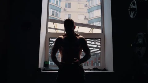The strong bodybuilder sits at the window with his chest bare, the ascending Stock Footage 156481128