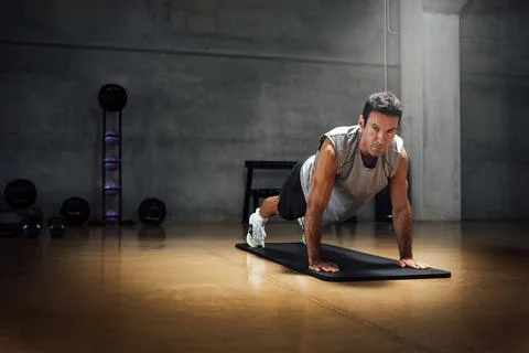 Strong caucasian man does push-ups while looks at camera in a dark gym. Stock Photos