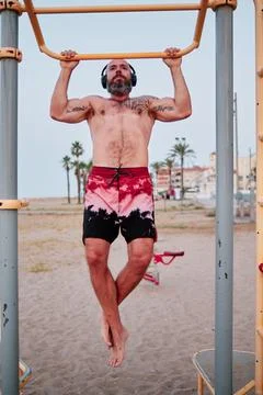 Strong man practicing calisthenics while doing barbell pull-ups on the beach. Stock Photos