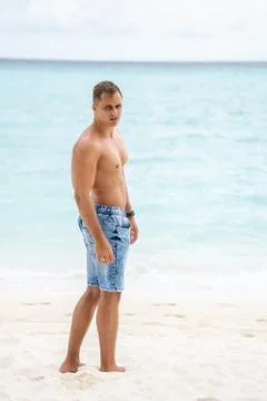 A strong young guy with a bare-chested walks on a white sandy beach on a trop Stock Photos
