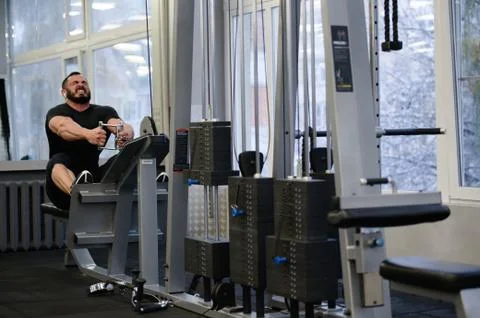 Strong young man pulling heavy weight sitting in sport fitness equipment indoors Foto stock