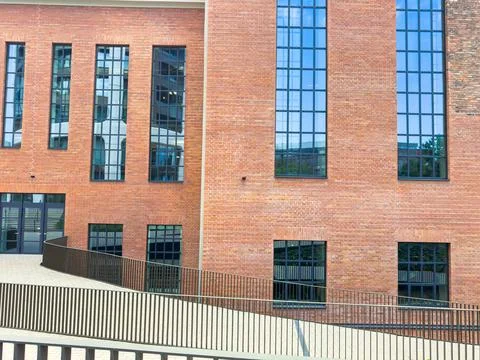 A structure made of red bricks with large glass windows. A sloped pathway lea Photos
