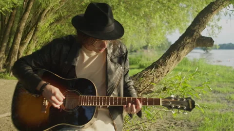 Strumming man guitar by the river under the trees at sunset Vídeos de archivo 319949081