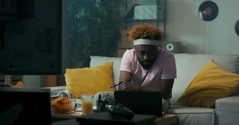 Student of African appearance with beard and curly hair is sitting on sofa. An 스톡 동영상 218091145