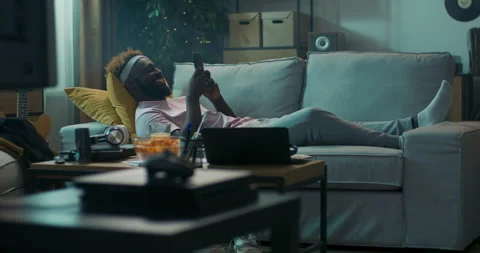 Student of African appearance is lying on sofa on yellow pillows, holding phone Vídeo Stock 218127674