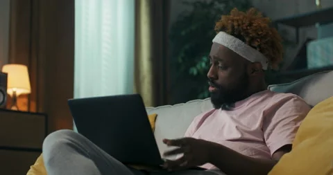 Student of African appearance is sitting on sofa, holding laptop and preparing 스톡 동영상 218100643