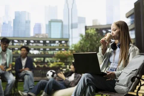 Student drinking coffee and working on laptop while seated on bench Stock Photos
