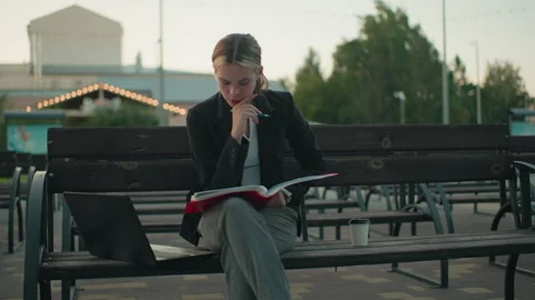 Student Focused Reading Book with Pen on Outdoor Bench Beside Laptop and Coffee Stock Footage 305654722