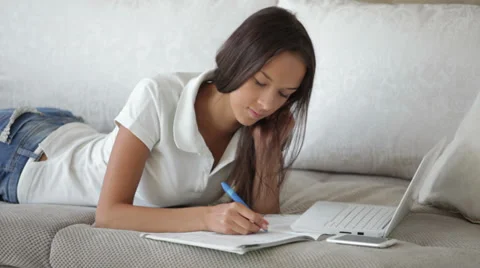 Student girl lying on sofa using laptop writing in workbook looking at camera Video stock 34196331