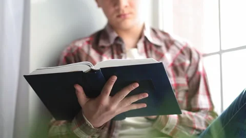 Student guy with a book in hands sits on a windowsill preparing for a lecture or Video stock 131867347