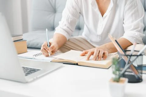Student hands writing notes while reading open book beside laptop on white .. 스톡 사진