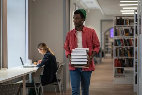 Student with huge stack of educational books goes to library workplace to Stock Photos