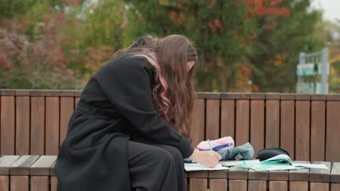 Student immersed in notetaking outside under cloudy sky. Female student in heavy Stock Footage 328145591