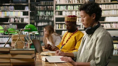 Student reading open book at library desk near chatbot on computer screen Stock Footage 337229208