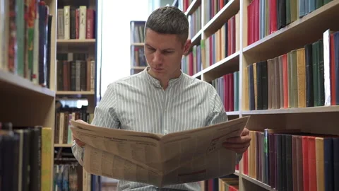 A student reads a newspaper between the rows of bookshelves in search of news. Stock Footage 219675860