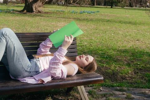 Student relaxing on a bench while reading a book outdoors on a sunny day Foto stock